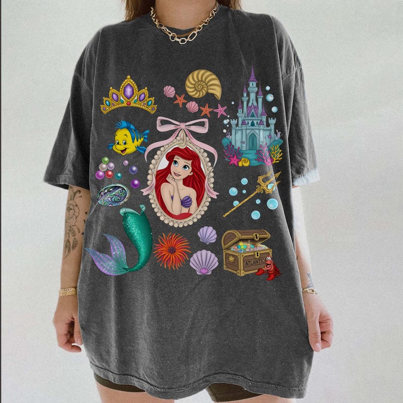 Puede incluir: Camiseta gris oscuro con un gr&aacute;fico colorido de Ariel de La Sirenita, una cola de sirena, un castillo y otras ilustraciones con tem&aacute;tica marina. El dise&ntilde;o incluye una tiara, conchas marinas y un cofre del tesoro. La camiseta tiene mangas cortas.