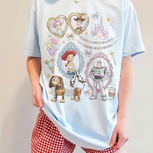 May include: Pale blue t-shirt with Toy Story cartoon characters. The design includes Woody, Buzz Lightyear, Jessie, and other characters in ornate frames, with the phrase "TO INFINITY AND BEYOND".