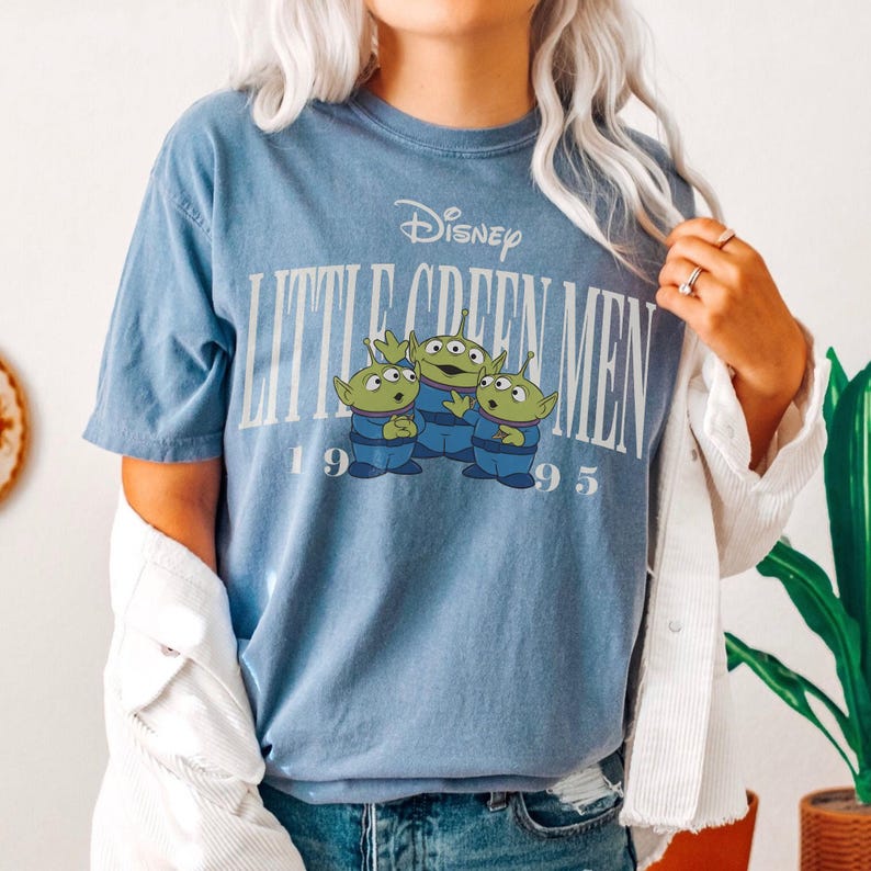 May include: A light blue t-shirt featuring the Disney logo and the words "Little Green Men" above a graphic of three green aliens. The year "1995" is printed below the aliens. The shirt has short sleeves and a crew neck.