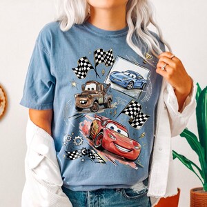 May include: Blue t-shirt featuring cartoon cars, including Lightning McQueen, Mater, and a blue race car. The design includes checkered flags, a trophy, and racing-themed graphics. The shirt is a light blue color.