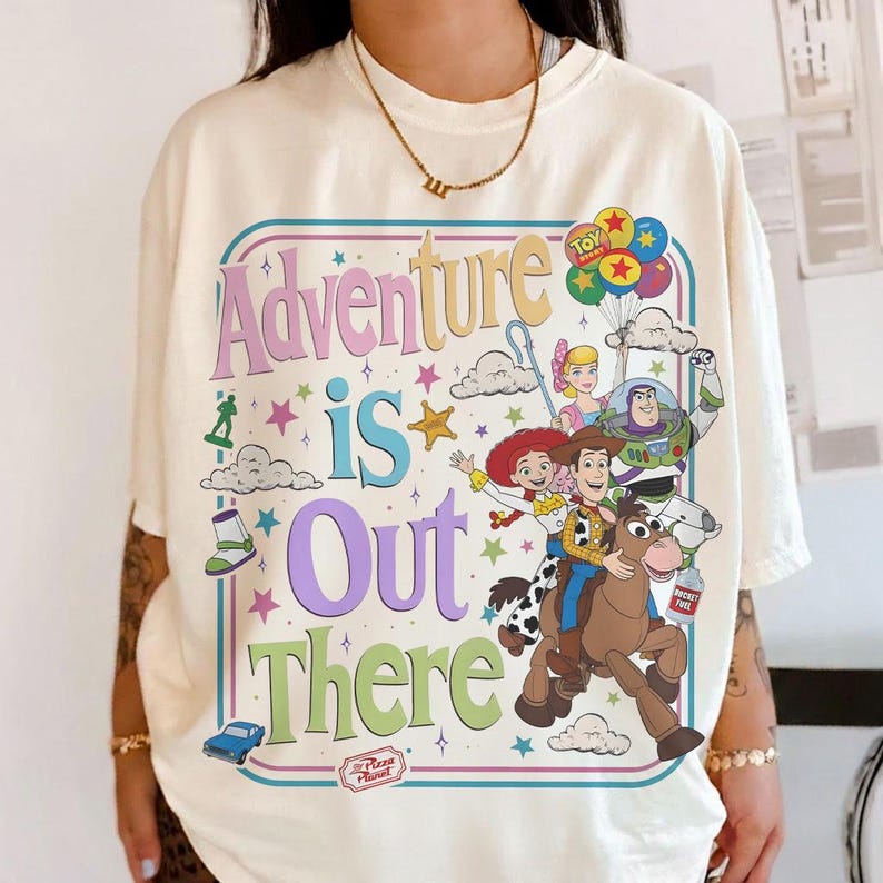 Pode incluir: Camiseta creme com um gr&aacute;fico colorido de personagens de Toy Story e a frase "Adventure is Out There". O design inclui Woody, Buzz Lightyear, Jessie e outros personagens, com estrelas e nuvens.