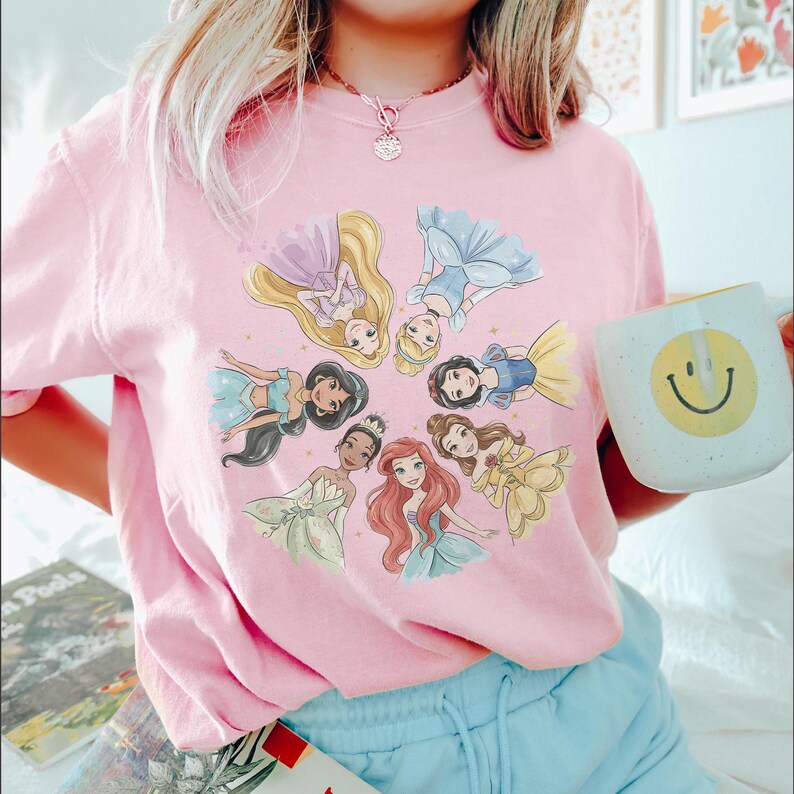 May include: Pink t-shirt featuring a circular graphic of animated princesses in various gowns. The princesses are surrounded by gold stars. A white mug with a yellow smiley face is held in the right hand.