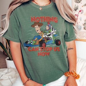 May include: Green t-shirt featuring a graphic of Woody and Buzz Lightyear in a race car, with the text "NOTHING CAN STOP US NOW" in red. The shirt has short sleeves and a crew neck.