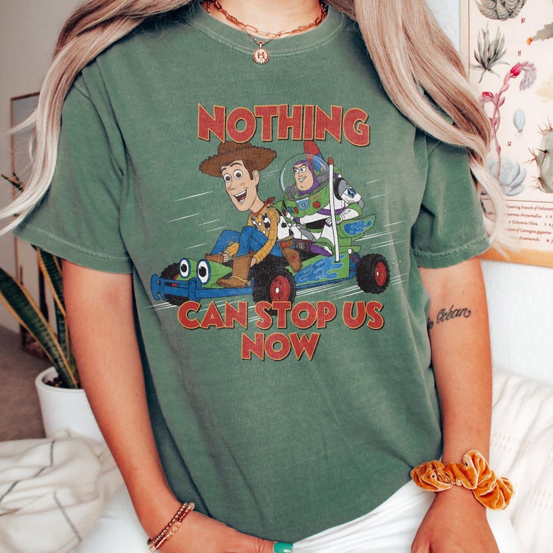 May include: Green t-shirt featuring a graphic of Woody and Buzz Lightyear in a race car, with the text "NOTHING CAN STOP US NOW" in red. The shirt has short sleeves and a crew neck.