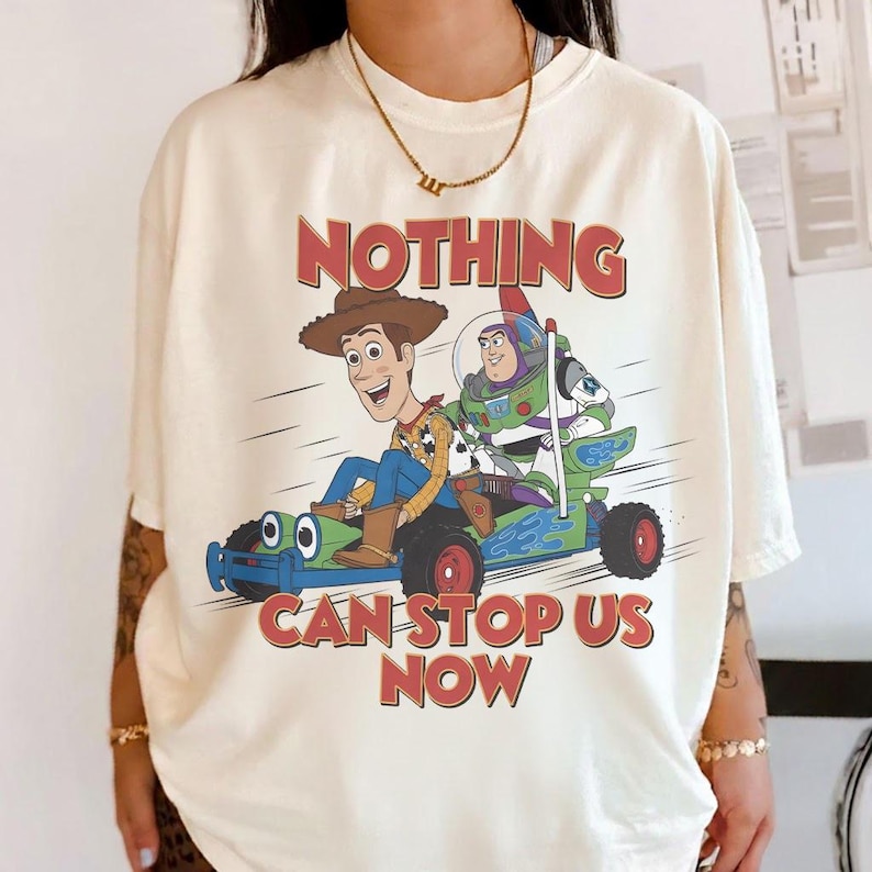 May include: Cream-colored t-shirt featuring a graphic of Woody and Buzz Lightyear from Toy Story in a race car. The text "NOTHING CAN STOP US NOW" is printed above and below the characters in red. The shirt has short sleeves and a crew neck.