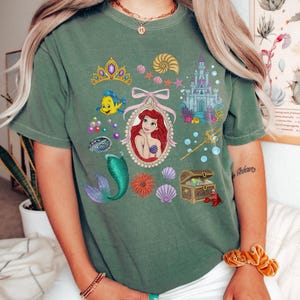 Puede incluir: Camiseta verde con un gr&aacute;fico colorido de Ariel de La Sirenita. El dise&ntilde;o incluye un retrato de Ariel, una cola de sirena, un castillo y otros elementos tem&aacute;ticos marinos. La camiseta es de un estilo c&oacute;modo e informal.