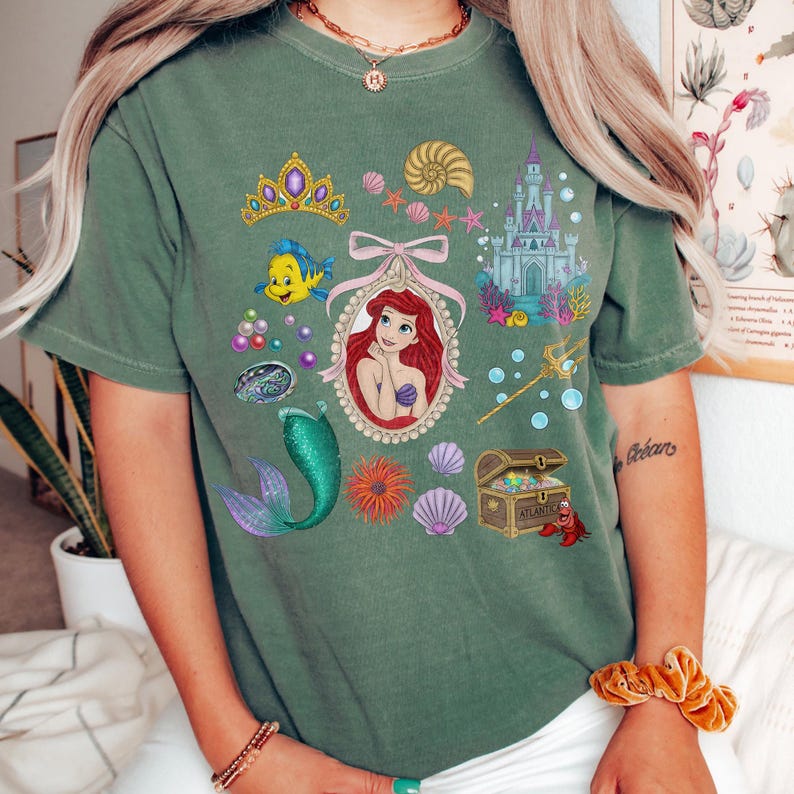 Puede incluir: Camiseta verde con un gr&aacute;fico colorido de Ariel de La Sirenita. El dise&ntilde;o incluye un retrato de Ariel, una cola de sirena, un castillo y otros elementos tem&aacute;ticos marinos. La camiseta es de un estilo c&oacute;modo e informal.