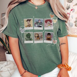May include: Sage green t-shirt featuring a graphic of Toy Story characters in polaroid-style frames. The design includes Woody, Buzz Lightyear, Jessie, Rex, Slinky Dog, and Mr. Potato Head. Mickey Mouse balloon graphics are also present.