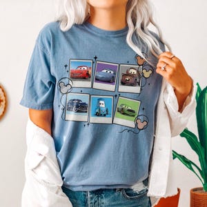May include: A light blue t-shirt featuring a graphic of six Cars movie characters in Polaroid-style frames. The design includes Mickey Mouse-shaped balloons. The shirt is made of a soft material.