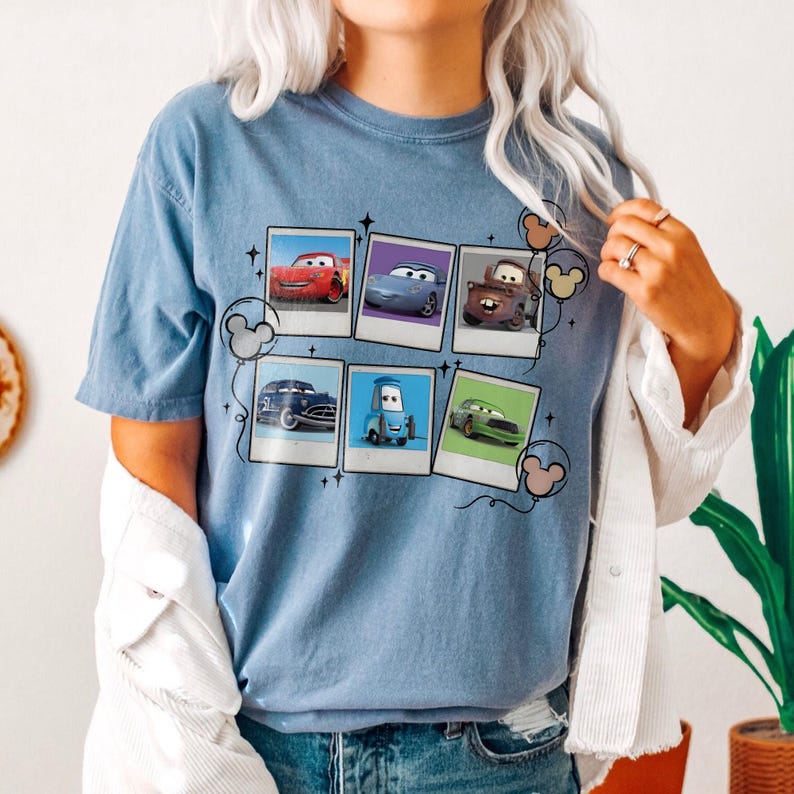 May include: A light blue t-shirt featuring a graphic of six Cars movie characters in Polaroid-style frames. The design includes Mickey Mouse-shaped balloons. The shirt is made of a soft material.
