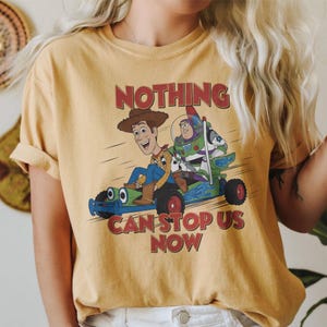 May include: Mustard yellow t-shirt featuring a cartoon graphic of Woody and Buzz Lightyear in a go-kart. The text "NOTHING CAN STOP US NOW" is printed in red. The shirt has short sleeves and a crew neck.