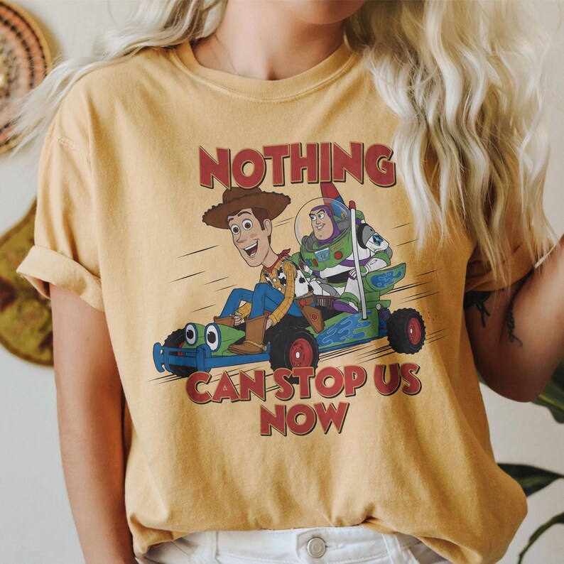 May include: Mustard yellow t-shirt featuring a cartoon graphic of Woody and Buzz Lightyear in a go-kart. The text "NOTHING CAN STOP US NOW" is printed in red. The shirt has short sleeves and a crew neck.
