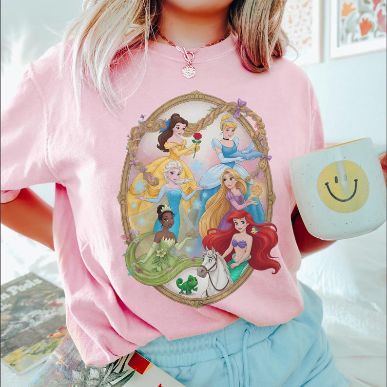May include: Pale pink t-shirt with a framed illustration of animated princesses. The princesses are in various gowns and poses. A white mug with a yellow smiley face is held in the right hand.
