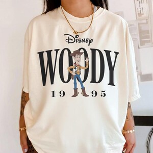 May include: Cream-colored t-shirt featuring the Disney logo and the name "WOODY" in large black letters. Below the name is the year "1995". A cartoon image of Woody, the Toy Story character, is in the center.