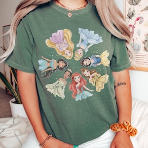 May include: Green t-shirt featuring a circular design of animated princesses. The princesses are depicted in various colors, including yellow, blue, pink, and green. The shirt is made of a soft material.