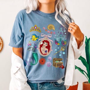 Puede incluir: Camiseta azul con un gr&aacute;fico colorido de una sirena, con una tiara, un castillo y otras ilustraciones con tem&aacute;tica marina. El dise&ntilde;o incluye un retrato de sirena, un cofre del tesoro y un tridente.