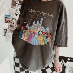 K&ouml;nnte beinhalten: Dunkelgraues T-Shirt mit einem farbenfrohen Aufdruck von Disney-Prinzessinnen vor einem Schloss, mit dem Schriftzug "Disney Princess" in Gold. Das Shirt hat kurze &Auml;rmel und eine lockere Passform.