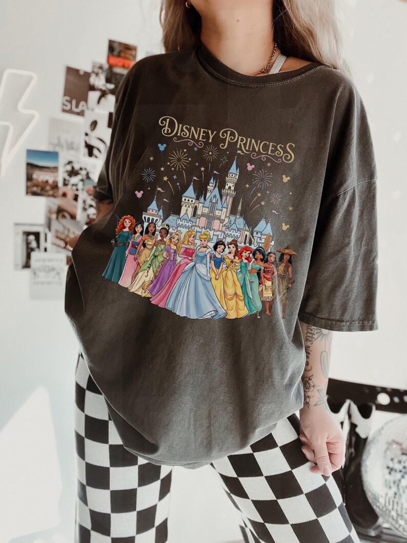 K&ouml;nnte beinhalten: Dunkelgraues T-Shirt mit einem farbenfrohen Aufdruck von Disney-Prinzessinnen vor einem Schloss, mit dem Schriftzug "Disney Princess" in Gold. Das Shirt hat kurze &Auml;rmel und eine lockere Passform.