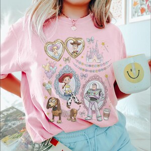 May include: A pink t-shirt with illustrations of Toy Story characters in decorative frames, such as Woody and Buzz Lightyear. The shirt also features a castle and the words "TO INFINITY AND BEYOND". A white mug with a yellow smiley face is held in the image.
