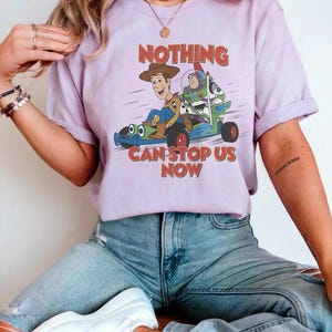 May include: A lilac-colored t-shirt featuring a graphic of Woody, Buzz Lightyear, and a dog in a race car. The text "NOTHING CAN STOP US NOW" is printed in red and blue. The shirt has short sleeves and a crew neck.