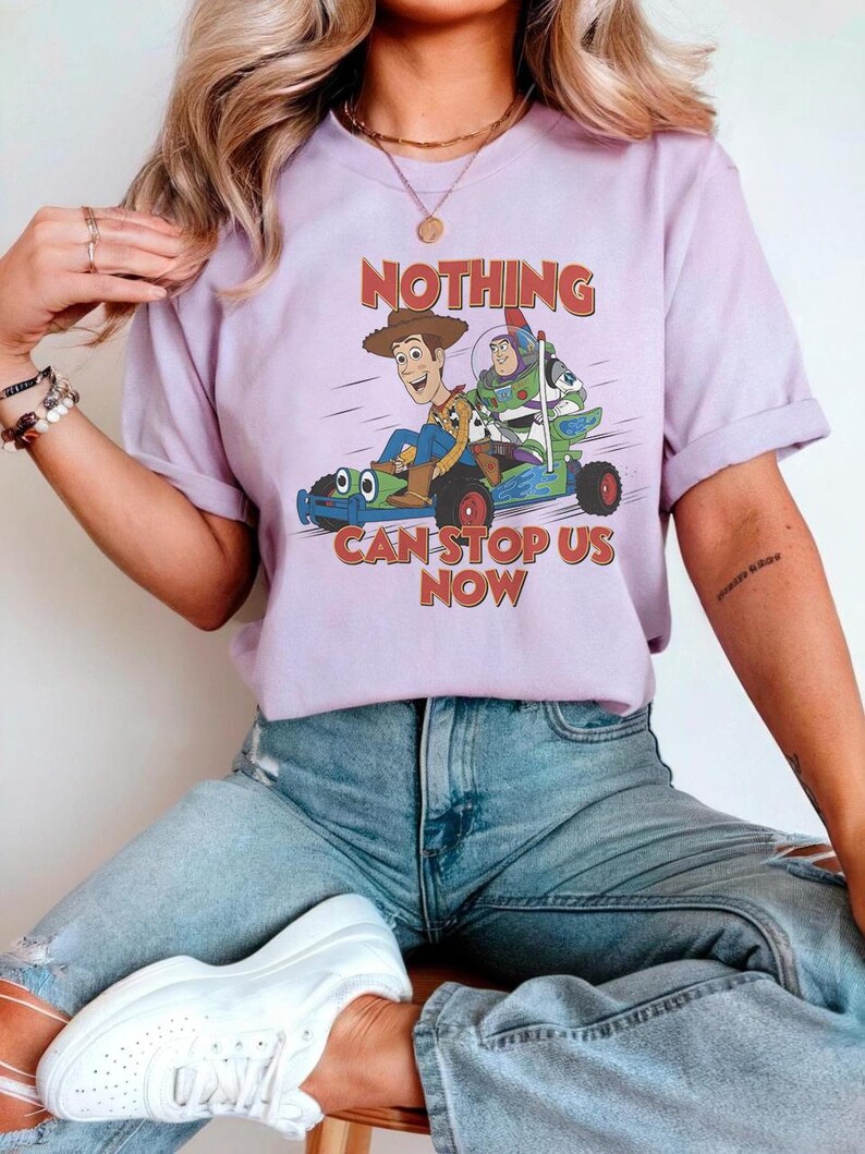 May include: A lilac-colored t-shirt featuring a graphic of Woody, Buzz Lightyear, and a dog in a race car. The text "NOTHING CAN STOP US NOW" is printed in red and blue. The shirt has short sleeves and a crew neck.
