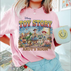 Puede incluir: Camiseta rosa claro con un gr&aacute;fico de Toy Story de Disney. El dise&ntilde;o incluye el t&iacute;tulo de la pel&iacute;cula, los personajes y el texto "Andy's Room". Se ve una taza blanca con una carita sonriente amarilla.