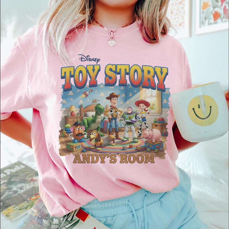 Puede incluir: Camiseta rosa claro con un gr&aacute;fico de Toy Story de Disney. El dise&ntilde;o incluye el t&iacute;tulo de la pel&iacute;cula, los personajes y el texto "Andy's Room". Se ve una taza blanca con una carita sonriente amarilla.