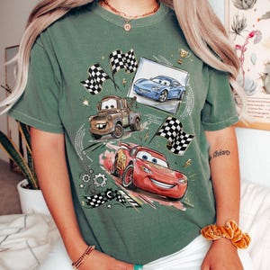 May include: Green t-shirt featuring a graphic of animated race cars, including Lightning McQueen, Mater, and a blue car, with checkered flags and a trophy. The design is centered on the shirt, with a vintage, slightly faded look.