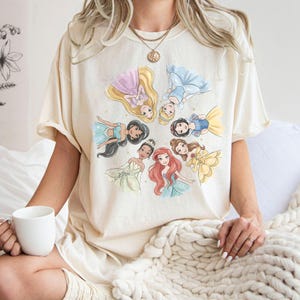 May include: Cream-colored t-shirt featuring a watercolor illustration of several princesses in a circular arrangement. The princesses are depicted in various gowns and hairstyles, with gold stars scattered around them. The shirt has short sleeves and a relaxed fit.