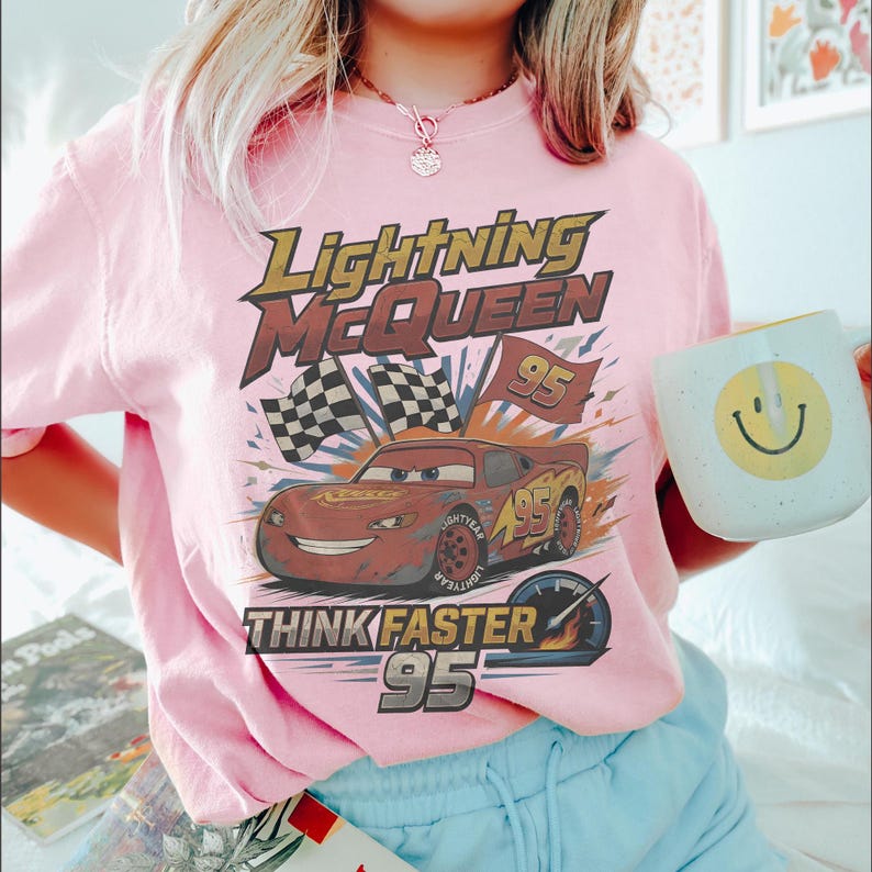 Pode incluir: Camiseta rosa claro com um desenho do Rel&acirc;mpago McQueen do filme Carros. O design inclui o texto "Lightning McQueen", "Think Faster" e o n&uacute;mero "95". Uma caneca branca com um rosto sorridente amarelo &eacute; segurada na imagem.