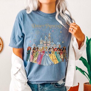 K&ouml;nnte beinhalten: Hellblaues T-Shirt mit dem Text "Disney Princess" &uuml;ber einer farbenfrohen Illustration eines Schlosses und mehrerer Disney-Prinzessinnen. Die Prinzessinnen tragen Kleider in verschiedenen Farben.