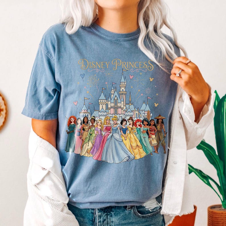 K&ouml;nnte beinhalten: Hellblaues T-Shirt mit dem Text "Disney Princess" &uuml;ber einer farbenfrohen Illustration eines Schlosses und mehrerer Disney-Prinzessinnen. Die Prinzessinnen tragen Kleider in verschiedenen Farben.