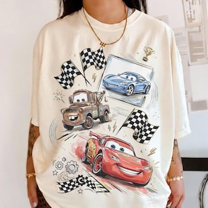 May include: Cream-colored t-shirt featuring cartoon images of race cars, including a red car with the number 95, a brown tow truck, and a blue sports car. Checkered flags and a trophy are also depicted.