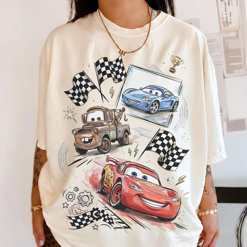 May include: Cream-colored t-shirt featuring cartoon images of race cars, including a red car with the number 95, a brown tow truck, and a blue sports car. Checkered flags and a trophy are also depicted.