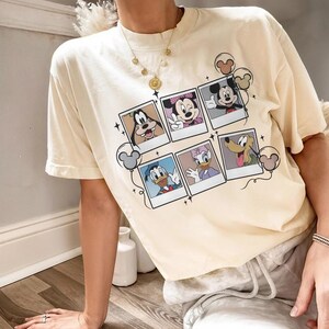 May include: Cream-colored t-shirt featuring a collage of Disney character polaroid-style images. The images include Mickey Mouse, Minnie Mouse, Donald Duck, Daisy Duck, Goofy, and Pluto. The shirt has a relaxed fit and short sleeves.