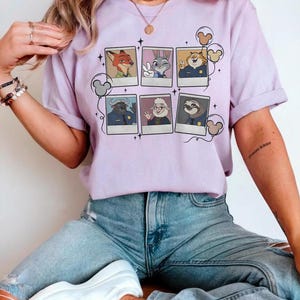 May include: A light purple t-shirt featuring a collage of Zootopia characters in polaroid-style frames. The design includes Nick Wilde, Judy Hopps, Chief Bogo, and others. The shirt has short sleeves and a relaxed fit.