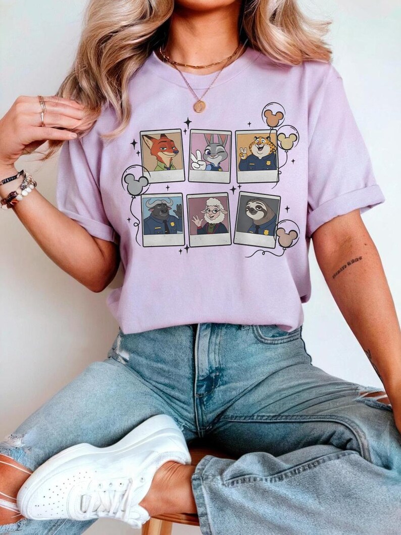 May include: A light purple t-shirt featuring a collage of Zootopia characters in polaroid-style frames. The design includes Nick Wilde, Judy Hopps, Chief Bogo, and others. The shirt has short sleeves and a relaxed fit.