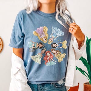 May include: Blue t-shirt featuring a circular design of cartoon princesses in various gowns. The princesses include characters with long blonde, brown, and red hair. The shirt is a solid blue color and has short sleeves.