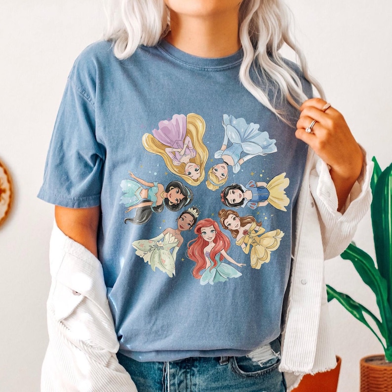 May include: Blue t-shirt featuring a circular design of cartoon princesses in various gowns. The princesses include characters with long blonde, brown, and red hair. The shirt is a solid blue color and has short sleeves.