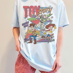 May include: Light blue t-shirt featuring a graphic of Woody, Buzz Lightyear, and Jessie from Toy Story. The shirt has the text "Toy Story" and "Since 1995" in a retro style. The shirt is worn with red and white checkered shorts.