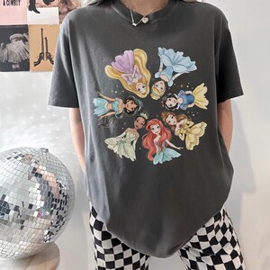 May include: Dark gray t-shirt featuring a circular design of cartoon princesses in various dresses. The princesses are in a circle, with their heads facing inward. The shirt is a casual style.