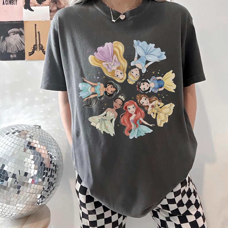 May include: Dark gray t-shirt featuring a circular design of cartoon princesses in various dresses. The princesses are in a circle, with their heads facing inward. The shirt is a casual style.