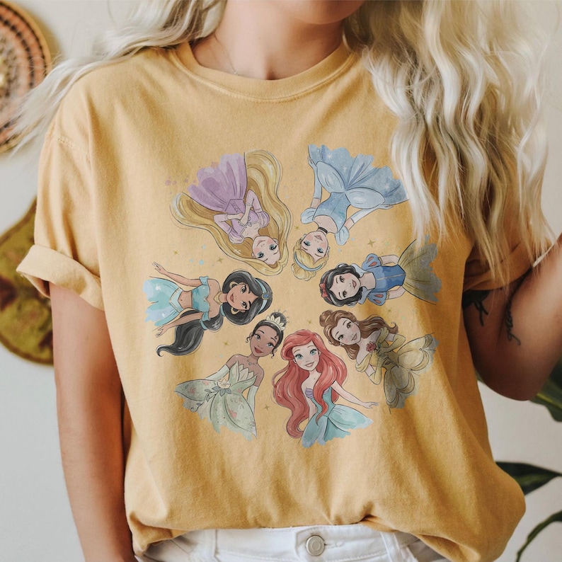 May include: Mustard yellow t-shirt featuring a circular design of animated princesses. The princesses are depicted in their signature gowns and hairstyles. The shirt has short sleeves and a crew neck.