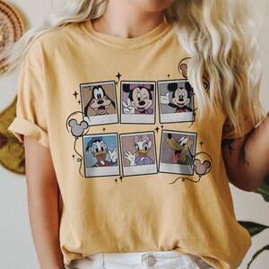 May include: Mustard yellow t-shirt featuring six cartoon character portraits in polaroid frames. The characters include Goofy, Minnie Mouse, Mickey Mouse, Donald Duck, Daisy Duck, and Pluto. The design includes Mickey Mouse ears and stars.