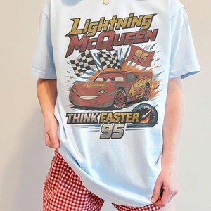 Pode incluir: Camiseta azul clara com Lightning McQueen do filme Carros. O design inclui o texto "Lightning McQueen", "Think Faster" e o n&uacute;mero "95". Apresenta tamb&eacute;m uma bandeira quadriculada e um veloc&iacute;metro.
