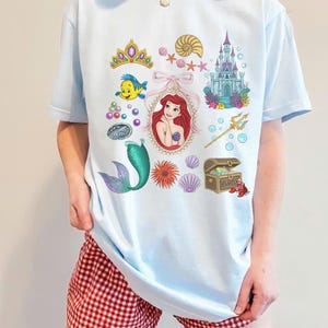 Puede incluir: Camiseta azul claro con ilustraciones coloridas de una sirena, un castillo, una corona y otros elementos marinos. La camiseta tiene cuello redondo y mangas cortas. El dise&ntilde;o es caprichoso y juguet&oacute;n.