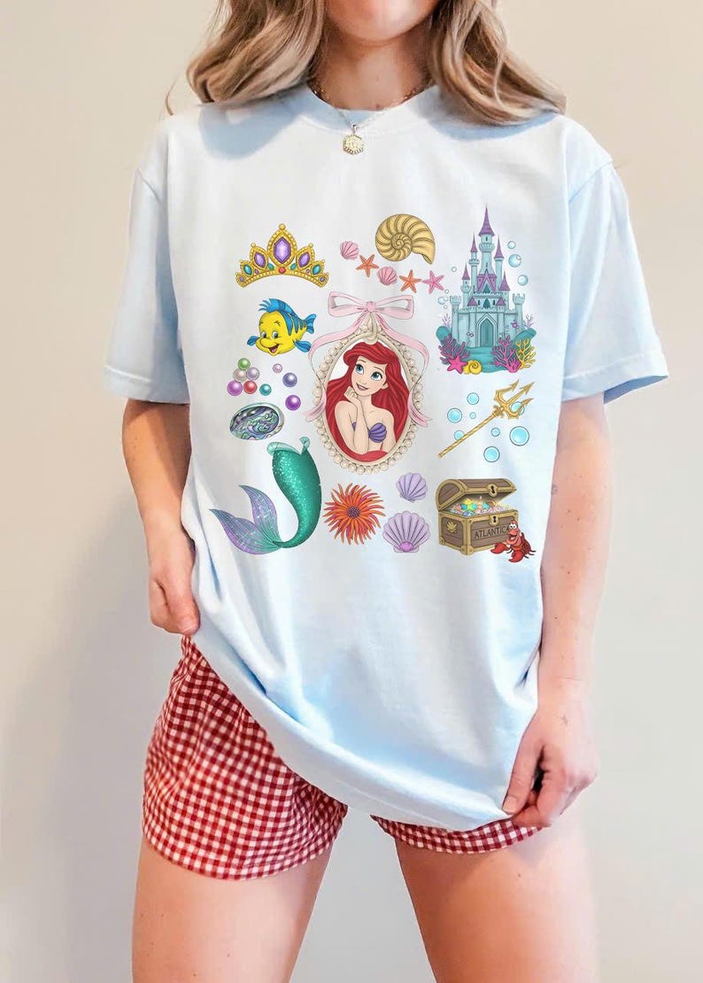 Puede incluir: Camiseta azul claro con ilustraciones coloridas de una sirena, un castillo, una corona y otros elementos marinos. La camiseta tiene cuello redondo y mangas cortas. El dise&ntilde;o es caprichoso y juguet&oacute;n.