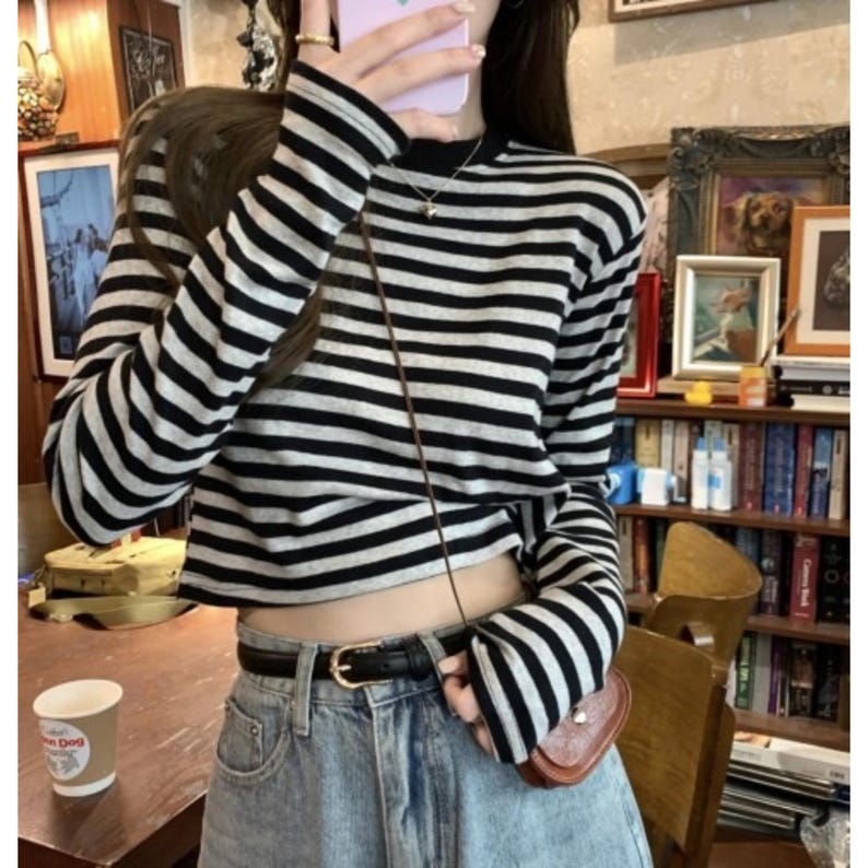 May include: A black and white striped long-sleeve crop top. The top is paired with light-wash denim jeans and a black belt with a gold buckle. A small brown purse hangs from a thin strap. A coffee cup with the word "Dog" is on the table.