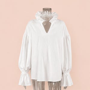 May include: A white blouse with a ruffled collar and a V-neckline. The long sleeves have a puffy design at the shoulders and end in ruffled cuffs. The blouse is made of a lightweight fabric.
