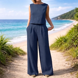 Könnte beinhalten: Ein marineblaues zweiteiliges Outfit. Das ärmellose Oberteil hat einen runden Ausschnitt. Die weite Hose hat Falten an der Taille und Taschen. Das Outfit ist aus leichtem Stoff gefertigt. Der Hintergrund zeigt einen Strand mit Meer und Himmel.